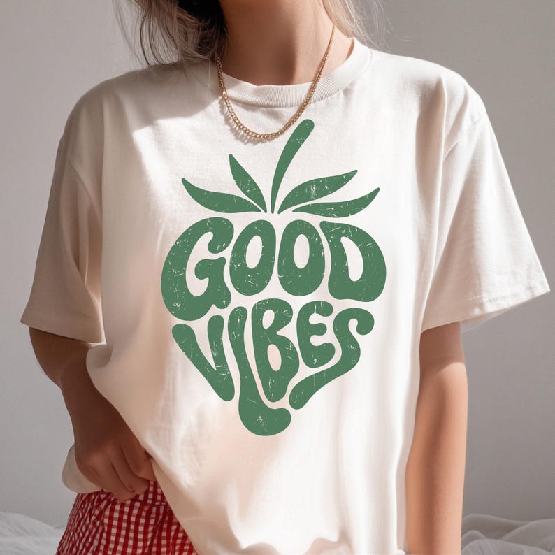 May include: Cream-colored t-shirt featuring a green graphic of a strawberry with the words "GOOD VIBES" in a retro font. The shirt has short sleeves and a crew neck. A gold chain necklace is visible.