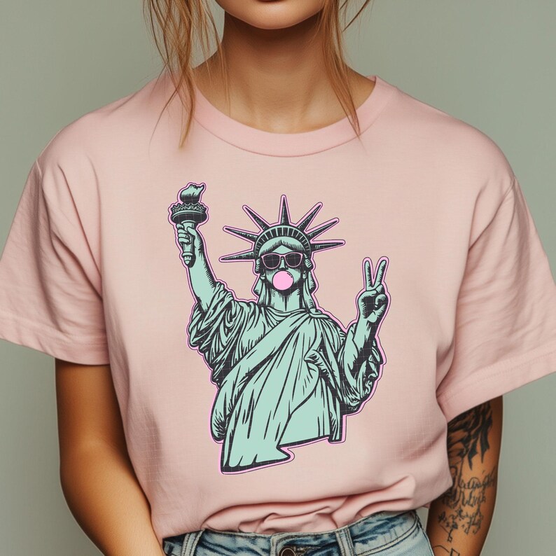 May include: A light pink t-shirt featuring a graphic of the Statue of Liberty wearing sunglasses and blowing a bubblegum bubble. The statue is holding a torch and making a peace sign. The graphic is outlined in black.