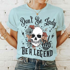 May include: A light blue t-shirt featuring a skull and floral design. The text "Don't Be Lady Be A Legend" is printed on the shirt.
