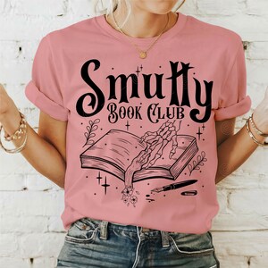 May include: A pink t-shirt with a black graphic design that says "Smutty Book Club". The graphic features a skeleton hand reaching into an open book with a quill pen and flowers.