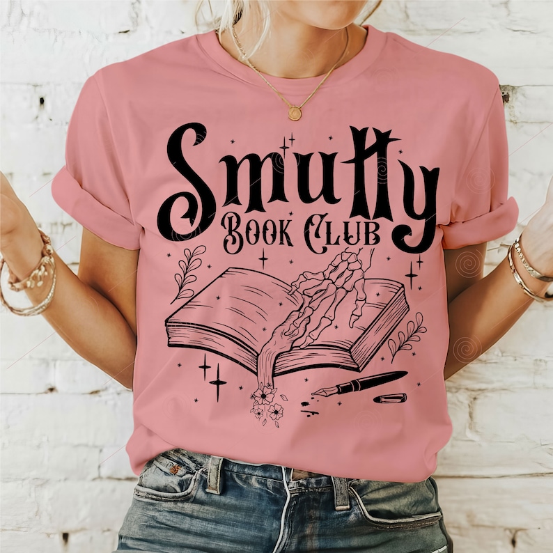 May include: A pink t-shirt with a black graphic design that says "Smutty Book Club". The graphic features a skeleton hand reaching into an open book with a quill pen and flowers.