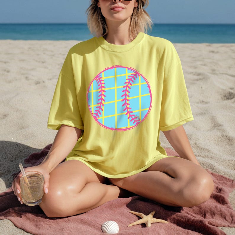 May include: A yellow t-shirt featuring a baseball graphic. The baseball design has a blue center with a yellow grid pattern and pink stitching. The shirt is worn on a beach setting.