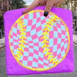 May include: A purple t-shirt featuring a baseball design. The baseball has a pink and light blue checkered pattern with yellow stitching and a yellow outline. The shirt is held up by a hand.
