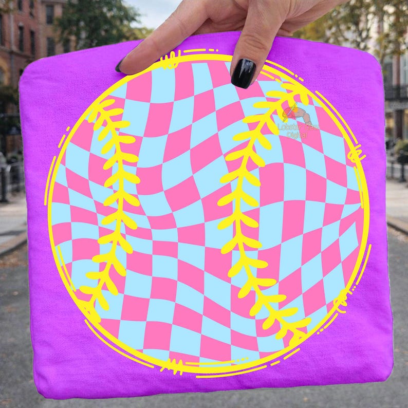 May include: A purple t-shirt featuring a baseball design. The baseball has a pink and light blue checkered pattern with yellow stitching and a yellow outline. The shirt is held up by a hand.