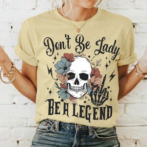 May include: A yellow t-shirt featuring a skull and flowers graphic. The text "Don't Be Lady Be A Legend" is printed on the shirt.