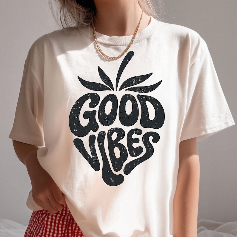 May include: Cream-colored t-shirt with a black graphic of a strawberry with the words "GOOD VIBES" in a retro font. The shirt has short sleeves and a crew neck. A gold chain necklace is visible.