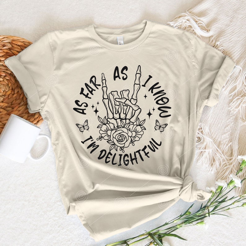 May include: A beige t-shirt with a black graphic print of a skeleton hand making a rock and roll sign. The graphic also includes flowers, butterflies, and the text "As far as I know I'm delightful".