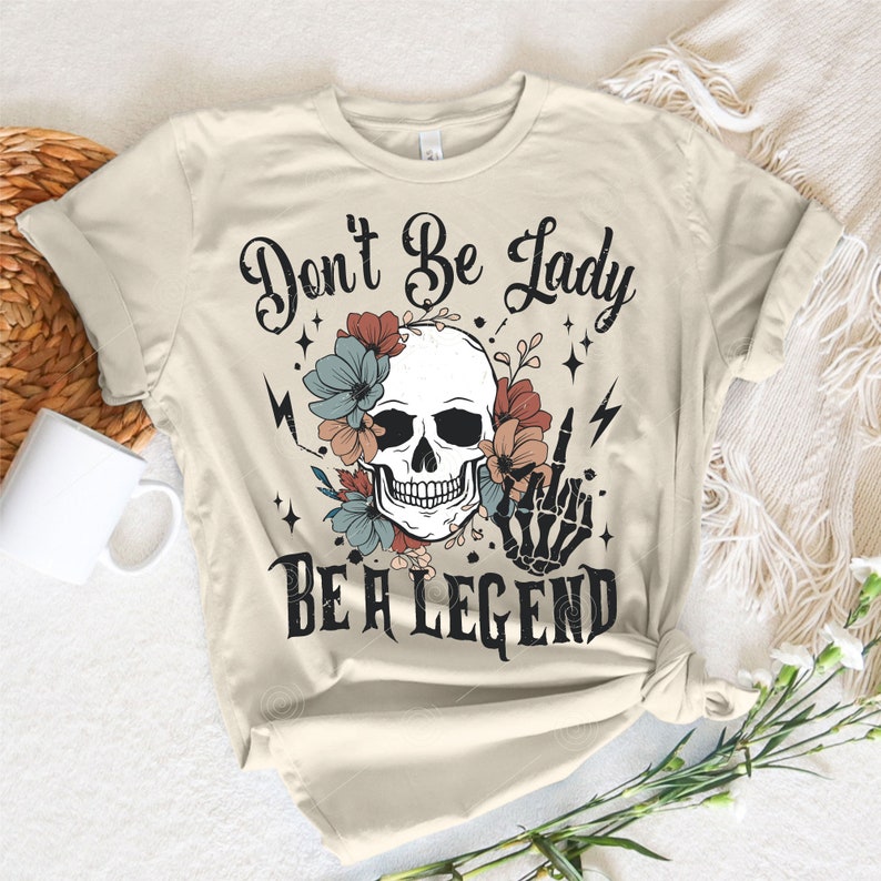 May include: A beige t-shirt featuring a skull and floral design. The text "Don't Be Lady Be A Legend" is printed on the shirt.