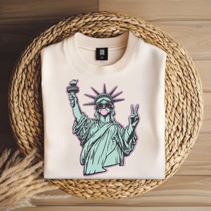May include: Cream-colored sweatshirt featuring a graphic of the Statue of Liberty wearing sunglasses and blowing a bubblegum bubble. The Statue of Liberty is holding a torch and making a peace sign. The sweatshirt is on a woven basket.