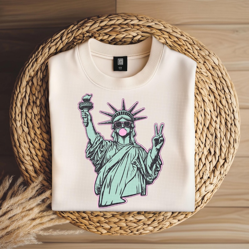May include: Cream-colored sweatshirt featuring a graphic of the Statue of Liberty wearing sunglasses and blowing a bubblegum bubble. The Statue of Liberty is holding a torch and making a peace sign. The sweatshirt is on a woven basket.
