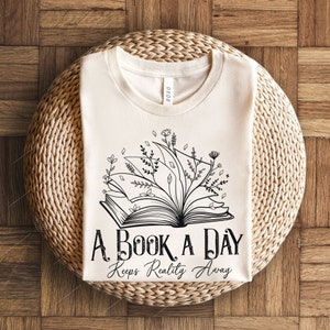 Op de afbeelding: Een wit T-shirt met een zwart-witte illustratie van een open boek met bloemen die eruit groeien. De tekst "A Book A Day Keeps Reality Away" is onder de illustratie gedrukt.