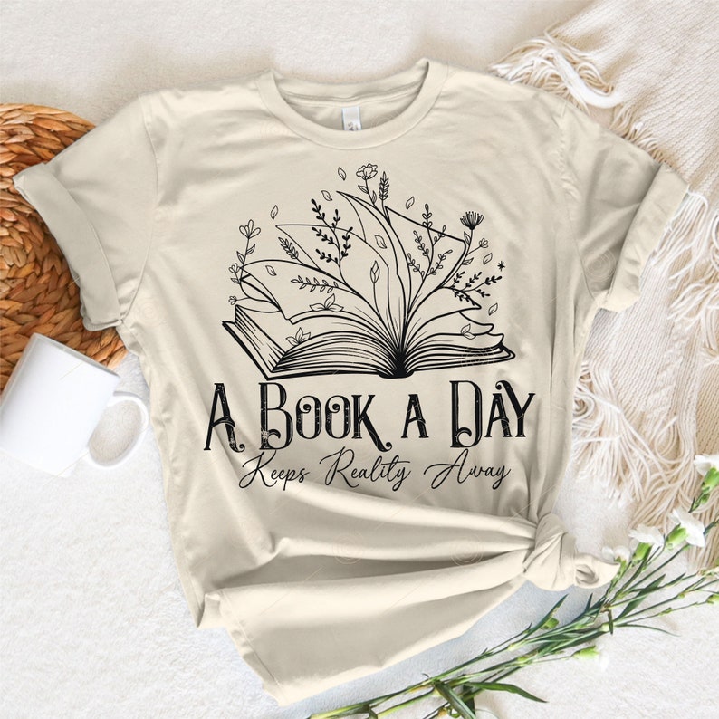 Op de afbeelding: Een beige T-shirt met een zwarte afbeelding van een open boek met bloemen die eruit groeien. De tekst "A Book A Day Keeps Reality Away" is onder het boek gedrukt.