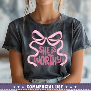 May include: A dark gray t-shirt with a pink bow and the text "She is worthy" printed on the front.