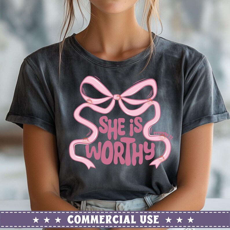 May include: A dark gray t-shirt with a pink bow and the text "She is worthy" printed on the front.