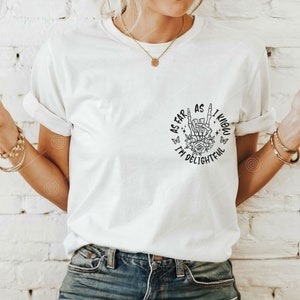 May include: White t-shirt with a black graphic print of a skeleton hand making a rock and roll sign with flowers and butterflies around it. The text reads "As far as I know I'm delightevil."