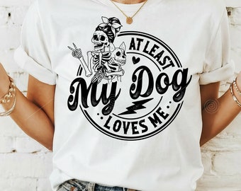 May include: White t-shirt with a black and white graphic design of a skeleton holding a dog. The graphic says "At Least My Dog Loves Me".