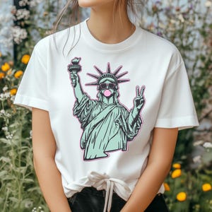 May include: White t-shirt featuring a graphic of the Statue of Liberty wearing sunglasses and blowing a bubblegum bubble. The Statue of Liberty is holding a torch and making a peace sign. The graphic has a teal and pink color scheme.