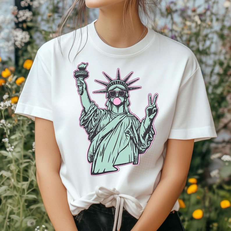May include: White t-shirt featuring a graphic of the Statue of Liberty wearing sunglasses and blowing a bubblegum bubble. The Statue of Liberty is holding a torch and making a peace sign. The graphic has a teal and pink color scheme.