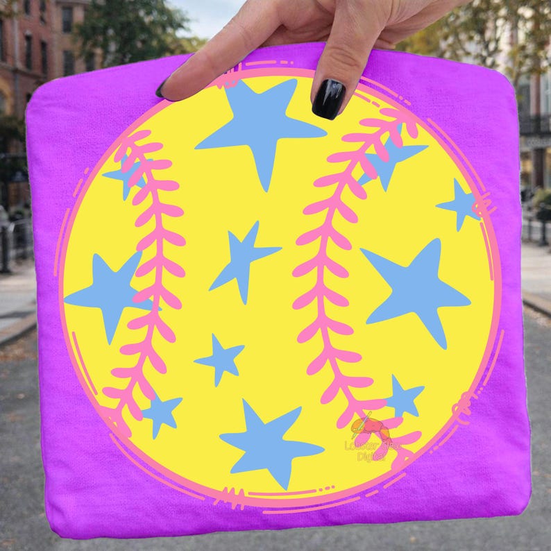 May include: A purple t-shirt featuring a softball design. The design includes a yellow circle with blue stars and pink leaf-like accents. The shirt is held up by a hand with a black painted fingernail.