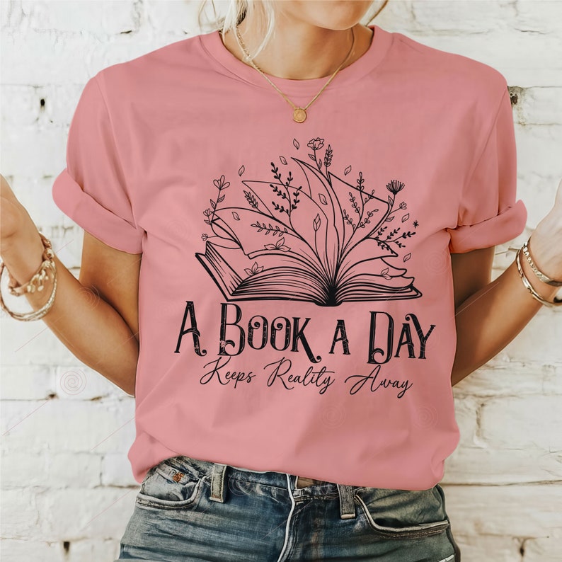 Op de afbeelding: Een roze T-shirt met een zwarte afbeelding van een open boek met bloemen die eruit groeien. De tekst "A Book A Day Keeps Reality Away" staat onder het boek.