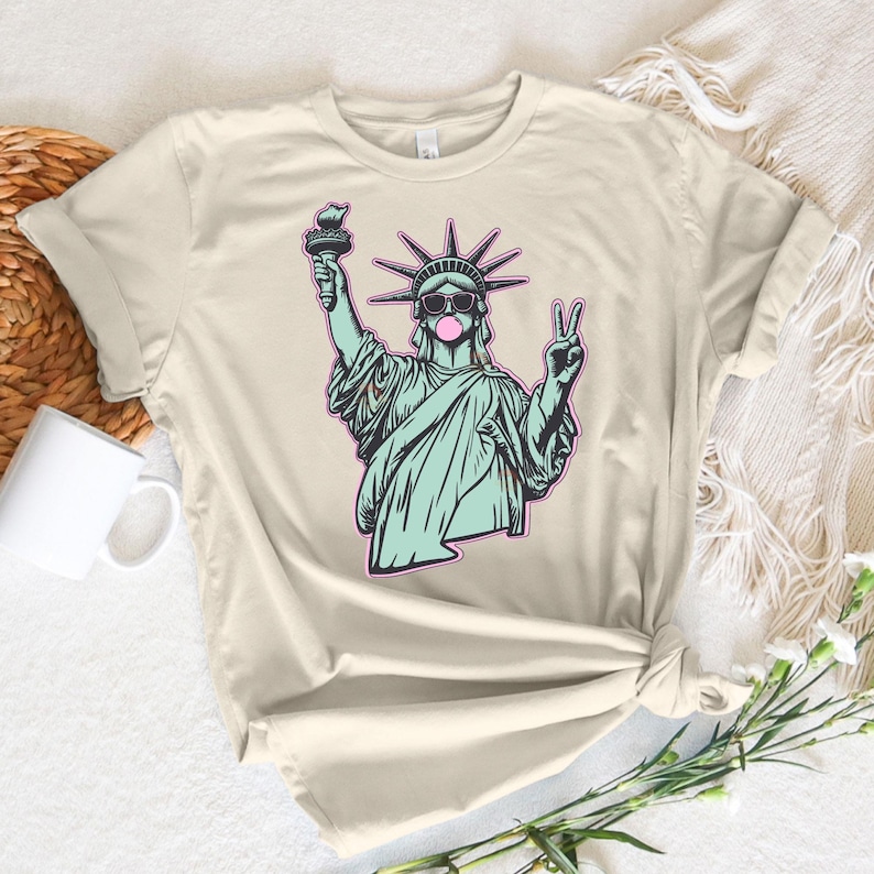 May include: Beige t-shirt featuring a graphic of the Statue of Liberty wearing sunglasses and blowing a bubblegum bubble. The statue is holding a torch and making a peace sign. The design is outlined in pink and teal.