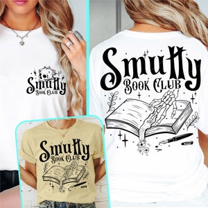 May include: Three white t-shirts with a black and white graphic design featuring a skeletal hand reaching out of an open book with the text "Smutty Book Club".