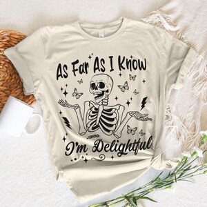 May include: A beige t-shirt with a black skeleton graphic and the text "As Far As I Know I'm Delightful". The skeleton is surrounded by butterflies and stars.