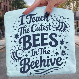 May include: A light blue tote bag with the text "I Teach The Cutest BEES In The Beehive" in dark blue. The design includes bee illustrations, hearts, and stars. The bag is held by a hand.