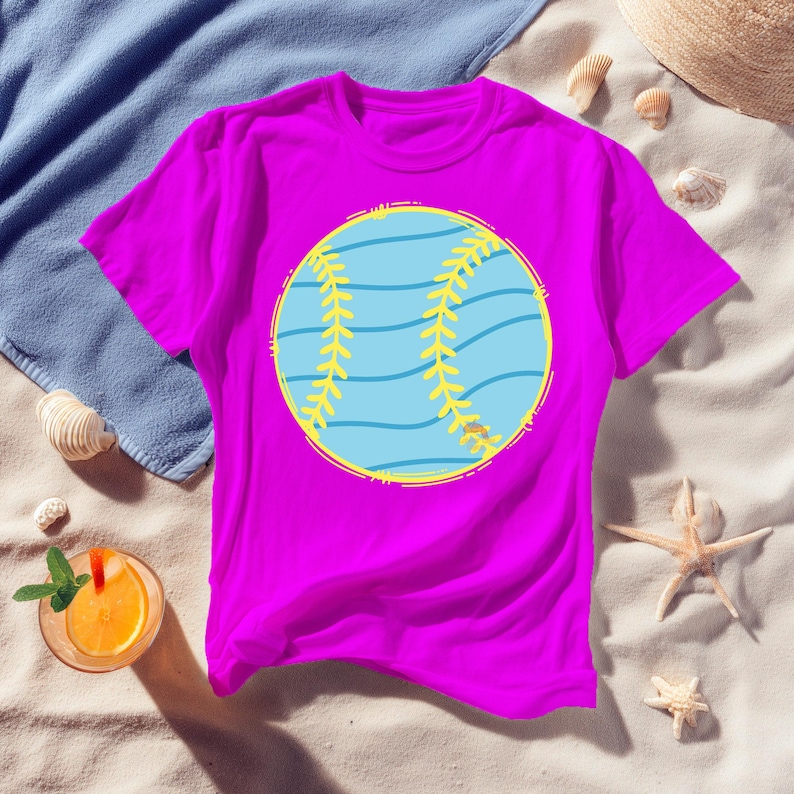 May include: A vibrant magenta t-shirt featuring a large, circular baseball graphic. The baseball design is light blue with yellow stitching and a yellow outline. The shirt is displayed on a sandy surface with beach-themed accessories.