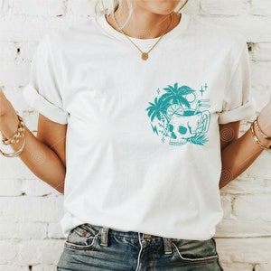 May include: White t-shirt with a teal blue graphic of a skull with a tropical drink inside. The skull is surrounded by palm trees, a lightning bolt, and stars.