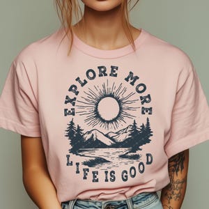 May include: Pink t-shirt with a graphic print of a sun, mountains, and a lake. The text "Explore More Life Is Good" is printed on the shirt.