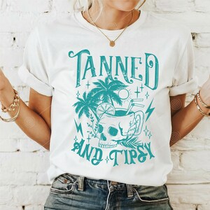 May include: White t-shirt with a teal graphic design that says "Tanned and Tipsy". The graphic features a skull in a mug with a lemon slice, palm trees, and stars.