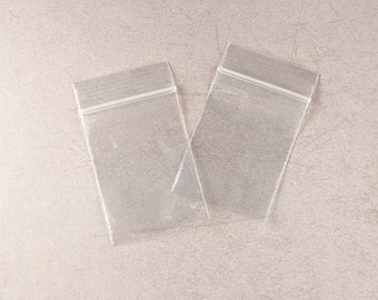 May include: Two clear, rectangular plastic bags with a zip-top closure are displayed on a neutral-toned surface. The bags are empty and transparent, showcasing their simple design and functionality. The zip-top is white.