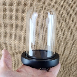 May include: A clear glass cloche with a rounded top, set on a black circular base. The cloche is empty, allowing for display of small items. The background is a textured tan fabric.