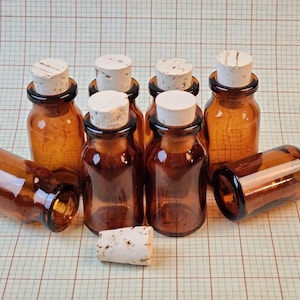 May include: A collection of small, amber-colored glass bottles with cork stoppers. The bottles are arranged on a grid-lined surface, some upright and some lying on their sides. The corks are white and cylindrical.