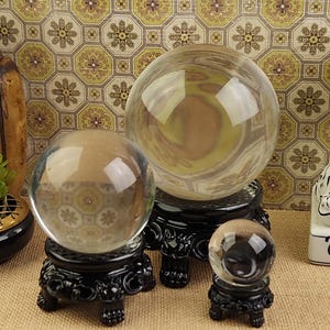 May include: Three clear crystal balls of varying sizes sit on ornate black stands. The largest ball has a yellow and brown interior. The stands have a detailed, carved design. The background features a patterned wallpaper.