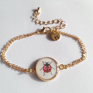 Könnte beinhalten: Goldkette Armband mit einem runden Anhänger mit einer roten und schwarzen Marienkäfer-Illustration. Das Armband hat einen kleinen Anhänger mit dem Buchstaben "M" darauf.