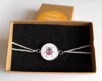 Puede incluir: Un collar de cadena de plata con un colgante redondo que presenta una mariquita pintada a mano sobre un fondo blanco. El collar se muestra en una caja marrón con un interior negro.
