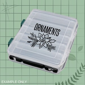 May include: Clear plastic storage box with a black label that says "ORNAMENTS" and a black silhouette of a poinsettia flower.