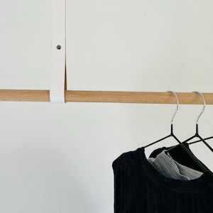 May include: A wooden clothes rod with a white leather strap hanging from the ceiling. Three black metal hangers are hanging from the rod with a black shirt on one of them.