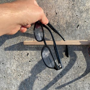 以下が含まれることがあります： 黒いフレームの眼鏡とべっ甲柄のサングラスが、水平な明るい色の木製スラットに掛けられています。眼鏡は質感のある灰色のコンクリートの表面に対して配置されており、フレームによって落とされた影がはっきりと見えています。