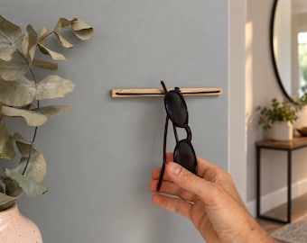 May include: A horizontal light-colored wooden wall-mounted rack featuring a thin black elastic cord stretched across its center. A pair of dark-rimmed sunglasses is suspended from the cord, hanging vertically against the wall surface.