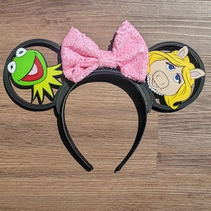 May include: Black headband with attached ears featuring Kermit the Frog and Miss Piggy. A large pink sequined bow sits between the ears. The characters are in a circular frame. The headband is on a wooden surface.