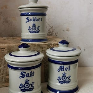 May include: Three white ceramic canisters with blue trim and lids. The canisters are labelled "Sukker", "Salt", and "Mel".