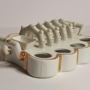 May include: A white porcelain inkwell with gold trim. The inkwell has a unique design with multiple pen holders and a small square ink pot. The piece is decorated with gold accents.