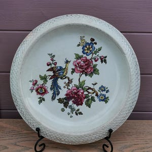 Peut inclure: Une assiette décorative en céramique blanche avec un bord texturé. L'assiette présente un motif floral et d'oiseaux coloré dans les tons de rose, de bleu et de jaune. L'assiette est présentée sur un support en métal noir.