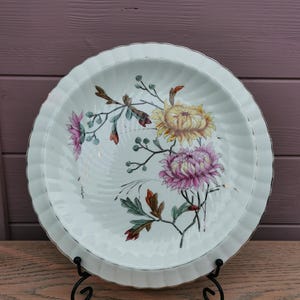 Peut inclure: Assiette en céramique blanche à bord festonné, ornée d'un motif floral avec des chrysanthèmes jaunes et roses, des feuilles vertes et des accents bruns. L'assiette est présentée sur un support en métal noir, sur fond de bois violet.