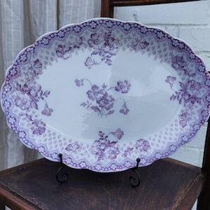 May include: An oval ceramic serving platter featuring a white base with a purple floral pattern. The rim is decorated with a scalloped edge and a detailed lace-like border. The centre displays a cluster of purple flowers and foliage.
