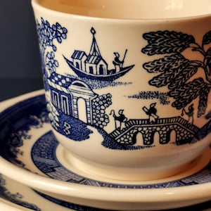 May include: A cream-colored teacup and saucer set with a blue Willow pattern. The cup features a detailed landscape scene with a boat, bridge, and buildings. The saucer has a matching border design. The set is made of ceramic.