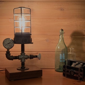 May include: A steampunk-style table lamp with a caged light bulb, a pressure gauge, and pipe fittings. The lamp is on a wooden base and is lit.
